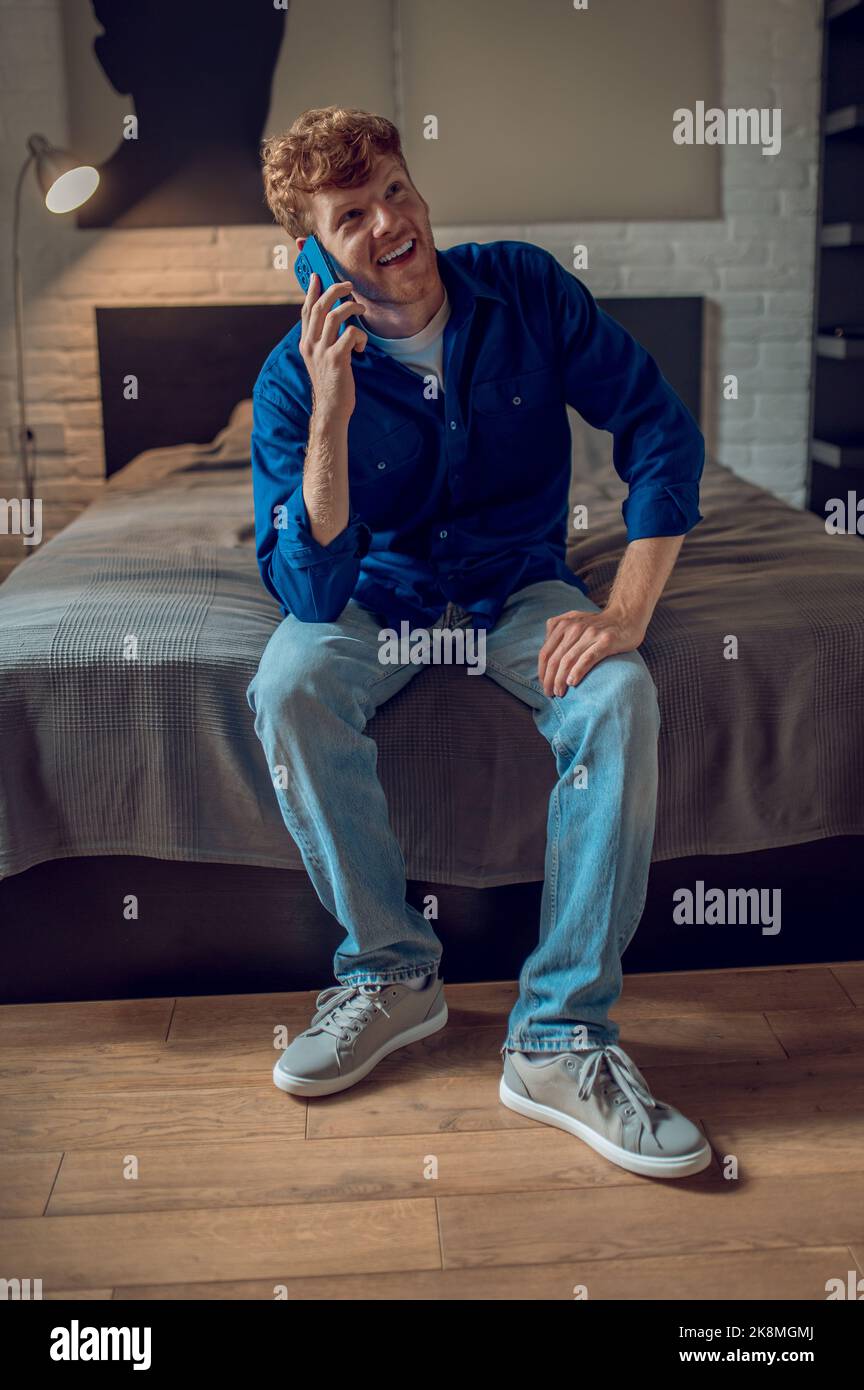 A man in dark-blue shirt sitting on the bed and talking on the phone Stock Photo