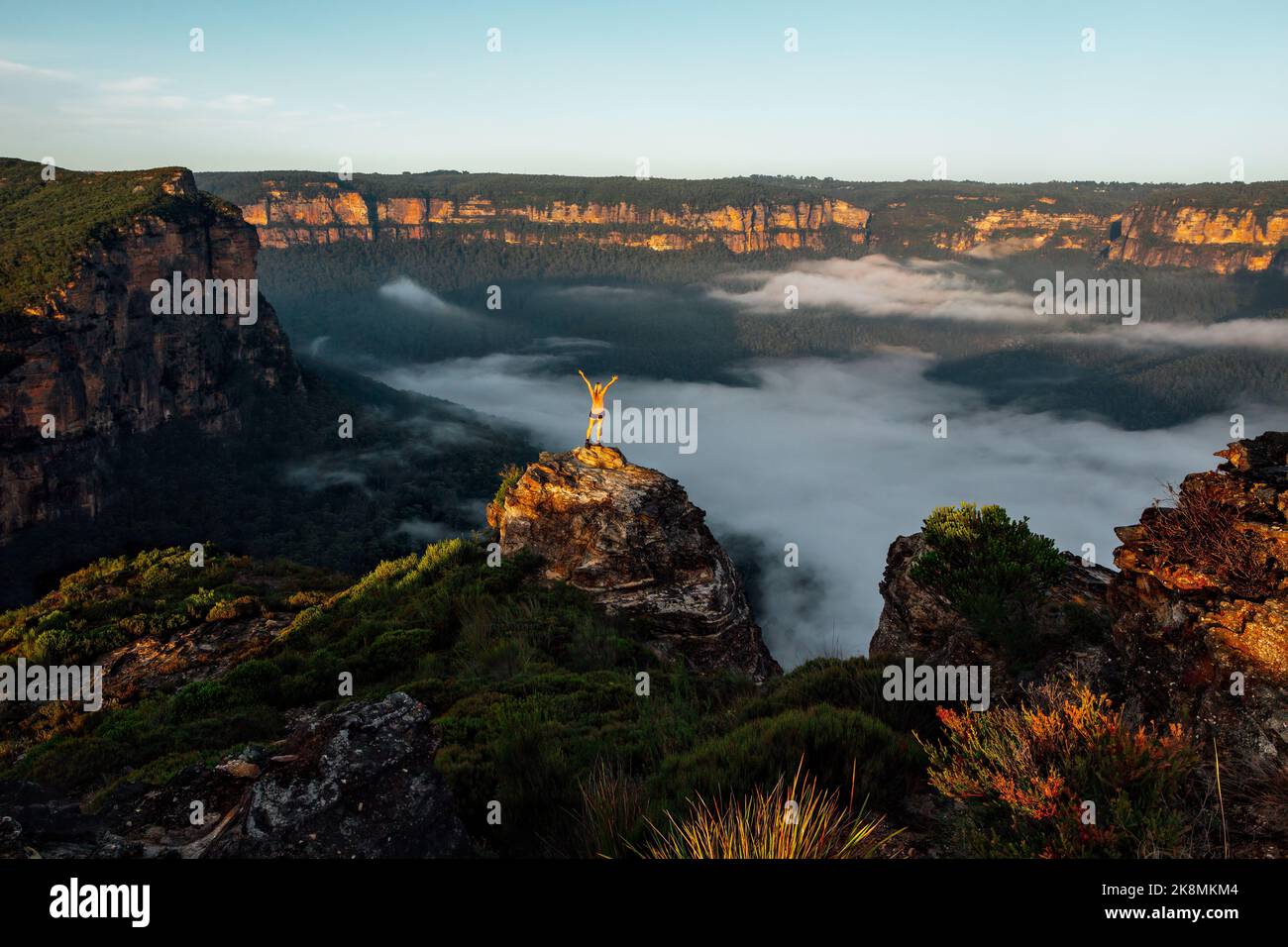 Woman hiker stands on a rocky pagoda ihigh in Blue Mountains  above a misty fog filled valley as first rays of sunlight hit the escarpment cliffs and Stock Photo
