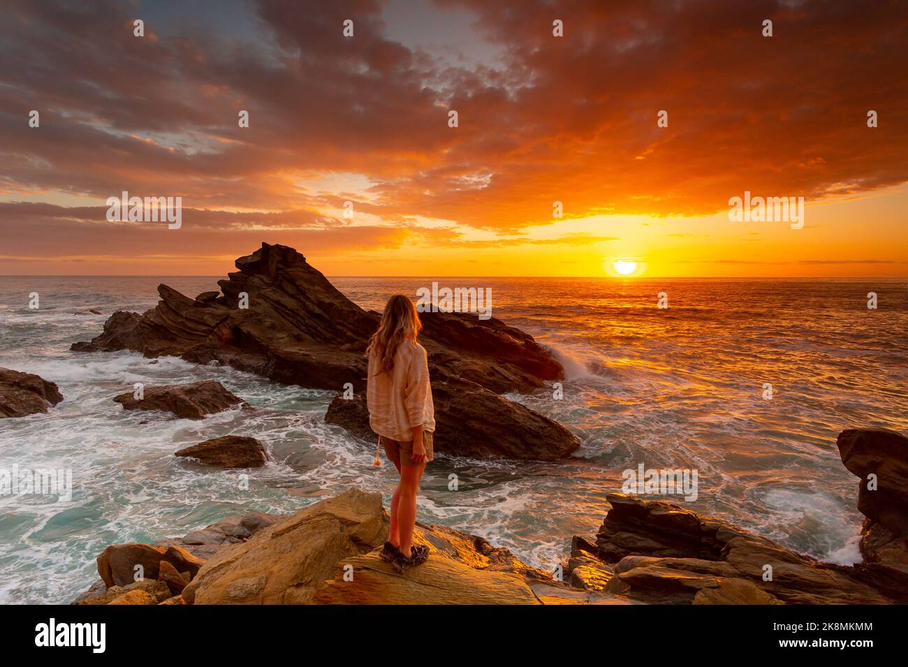 Woman wearing a casual cream linen shirt and shorts stands on a rock watching a vivid red sunrise over the ocean as waves crrash around her Stock Photo