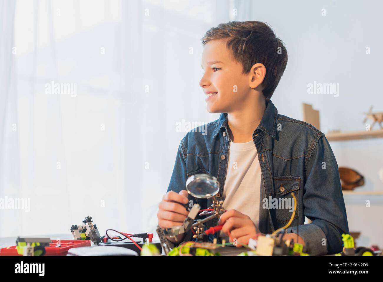 Smiling preteen boy holding magnifying glass near robotics model at home,stock image Stock Photo