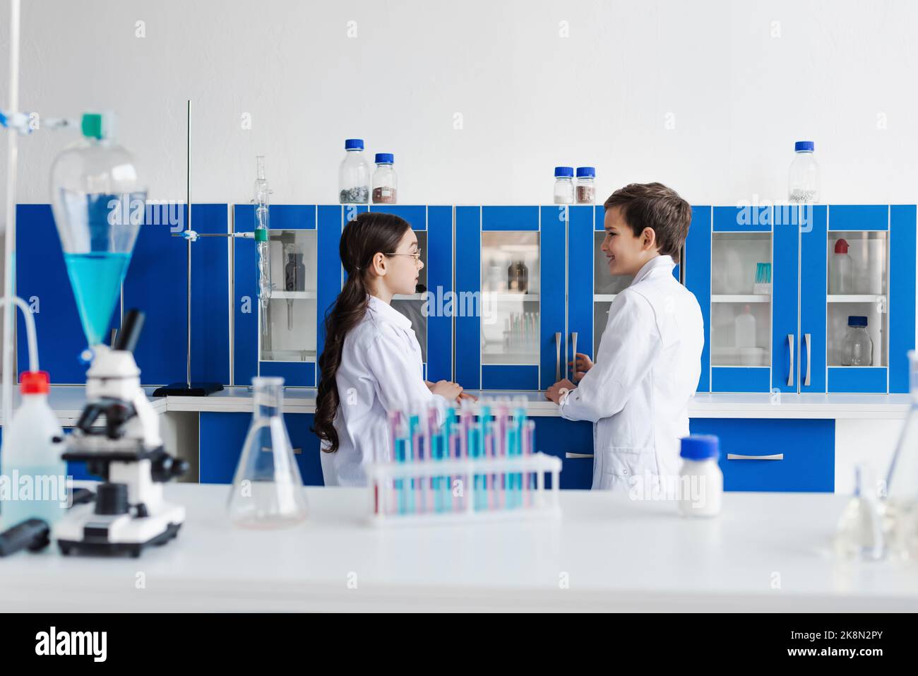 side view of kids in white coats talking near test tubes and microscope on desk,stock image Stock Photo
