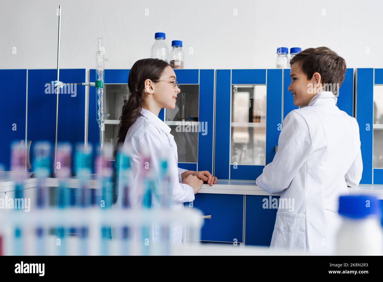 smiling friends talking near locker and test tubes on blurred foreground in lab,stock image Stock Photo