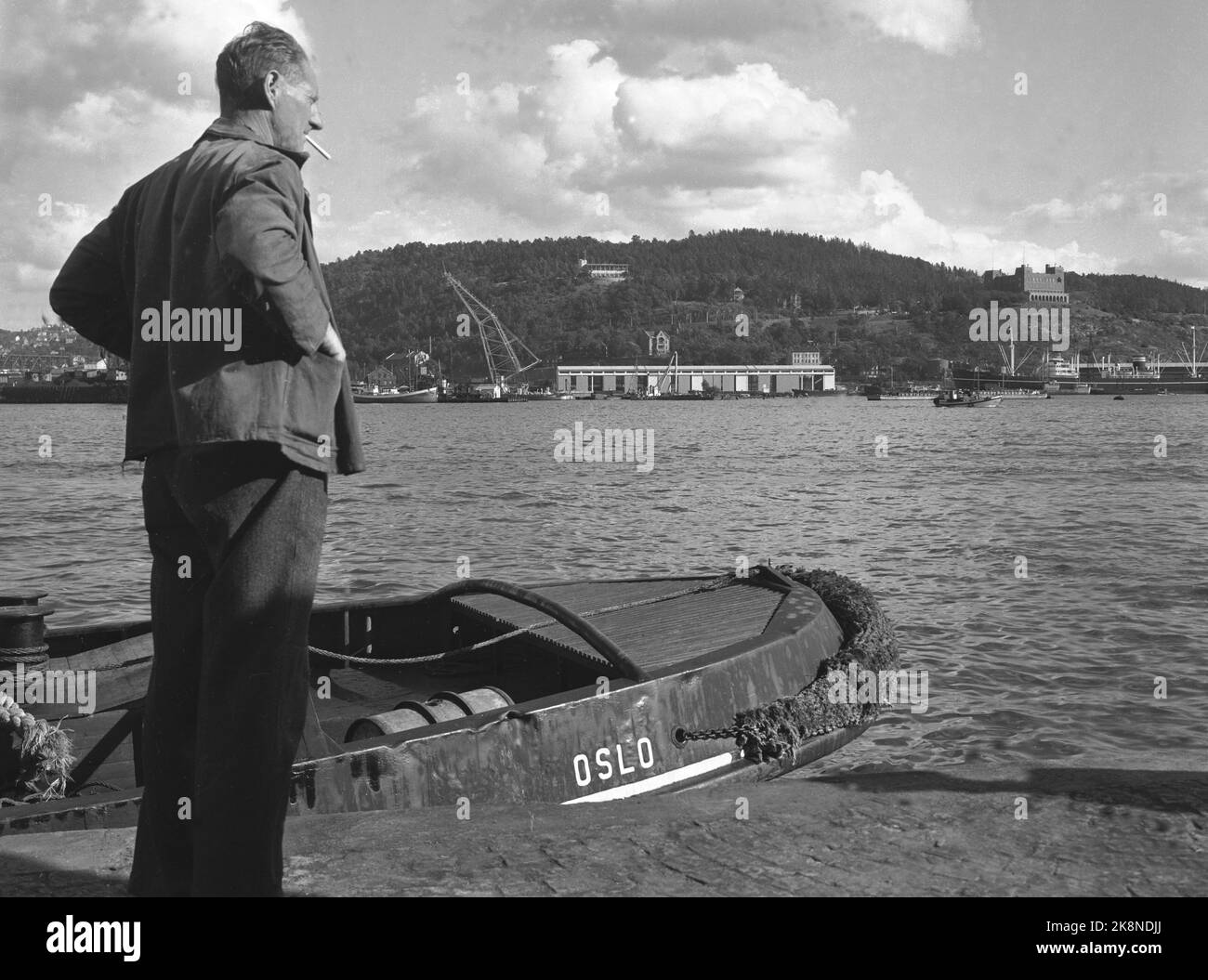 Oslo. 19490715. A man stands on the rocker with a cigarette in his mouth. In the background are Ekebergåsen and Bekkelaget with the Seamen's School T.H and the smaller Ekebergrestaurant t. v. In the picture. Grønlikaia with large warehouse and operating hall. Cranes and different types of boats also characterize the subject. Photo: NTB Stock Photo