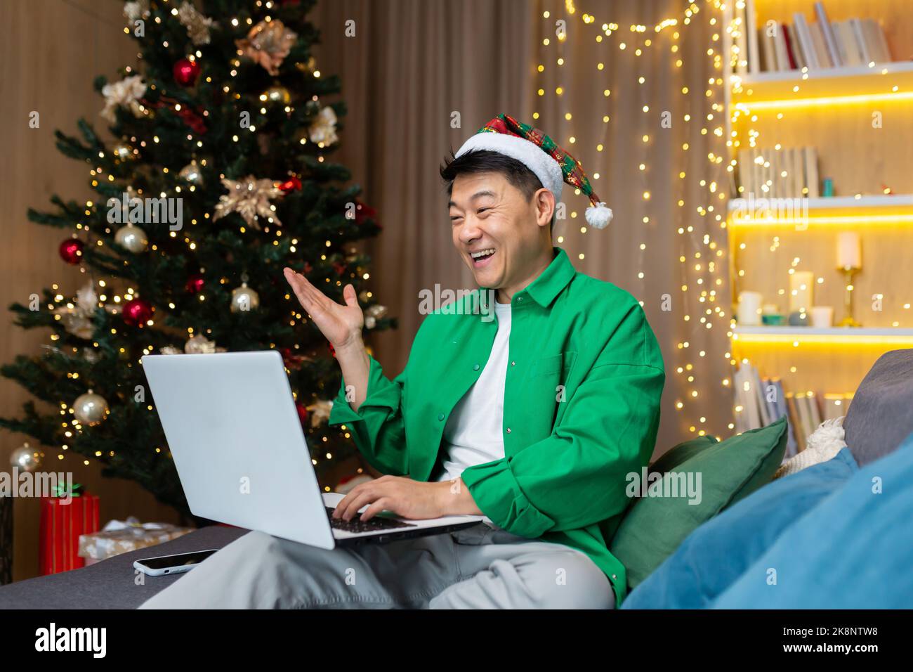 Cheerful man in christmas hat and green shirt talking with friends on video call, asian man celebrating christmas and new year at home sitting on sofa in living room with laptop. Stock Photo