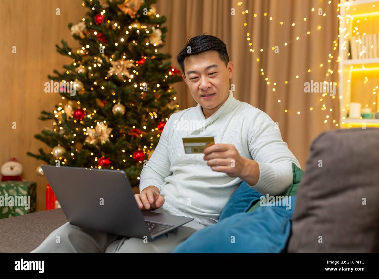 Successful businessman on christmas at home sitting on sofa in living room, using laptop online shopping, asian celebrating new year near tree, choosing gifts in online store shopping credit card. Stock Photo