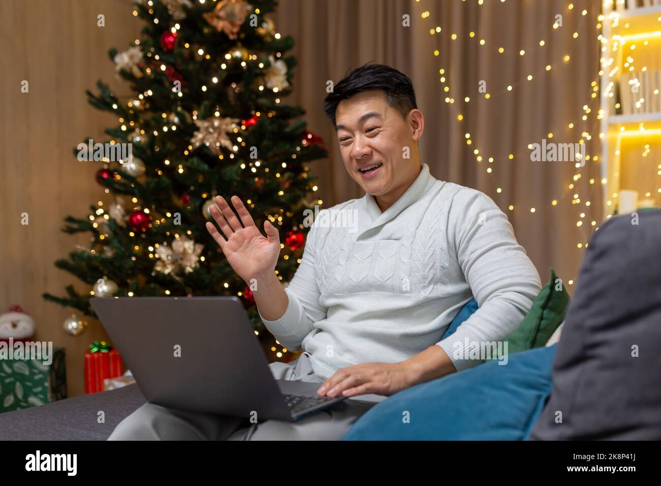 Asian man on Christmas at home talking on video call sitting on sofa in living room near Christmas tree, man celebrating new year waving at camera greeting gesture. Stock Photo