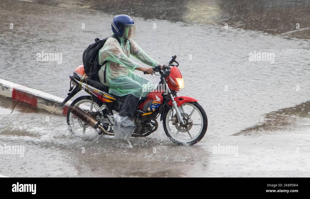 SAMUT PRAKAN, THAILAND, OCT 03 2022, A motorcyclist in a raincoat drives through a puddle in the rain Stock Photo