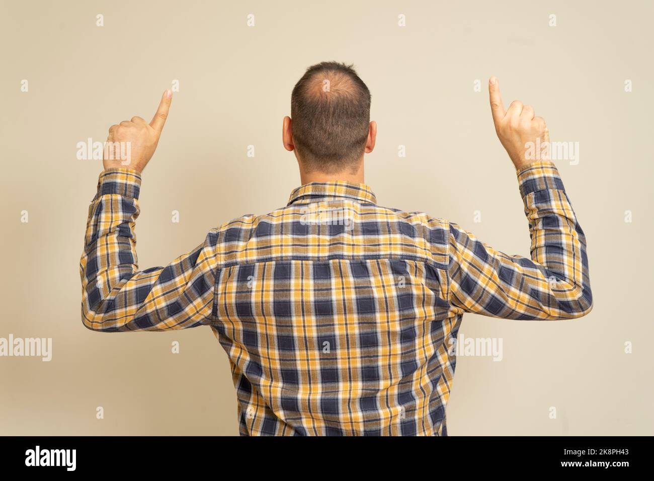 Portrait of a man with incipient alopecia from behind, raising both hands and pointing his index fingers upwards, isolated on a beige background. Stock Photo