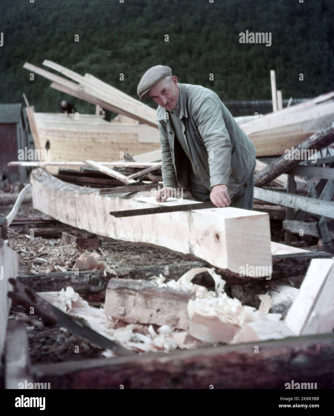 Rognan innerst i Saltdalsfjorden 19531010. Rognan er kjent over hele Nord-Norge som stedet hvor fiskeskøyter bygges og repareres. Småskipsbyggingen har urgamle tradisjoner, for hundrevis av år siden sto folk og bygde båter i små skur nede i fjæra. De fleste bøndene har en båt for seg selv på låven i vedskjulet eller i et skur. Båtene bygges i mørke høst--og vinterdager når arbeidet med gardsbruket ikke stjeler så mange timer av døgnet. Langs hele stranda i Rognan er skøyter under bygging eller til reparasjon. Her ser vi Harald Bakken høvler til en spant-planke.  Foto: Sverre A. Børretzen / Akt Stock Photo