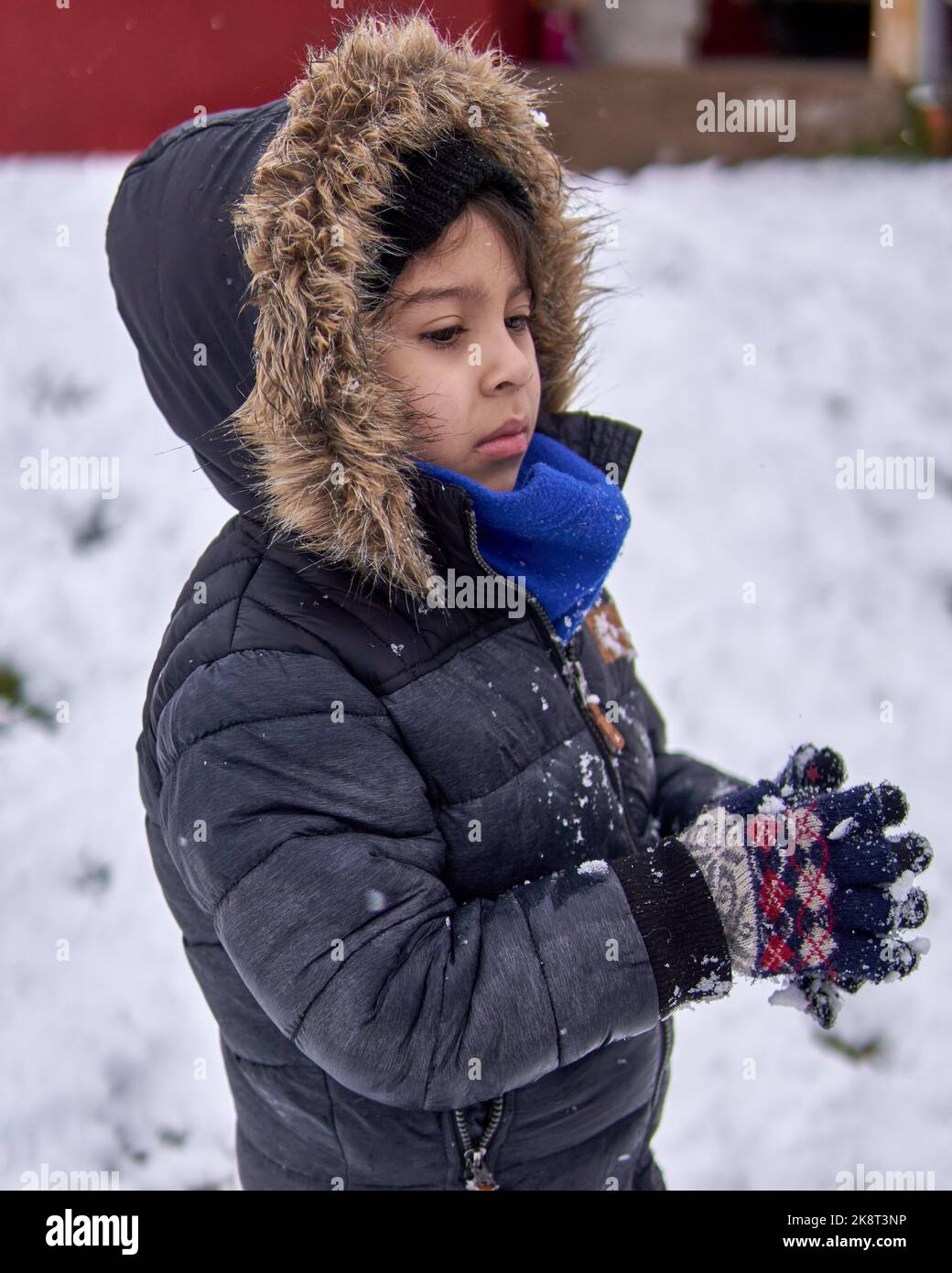 portrait of Latino little boy in profile with worried face and clasping his gloved hands together, in winter in the snow, wearing a furry hooded coat Stock Photo