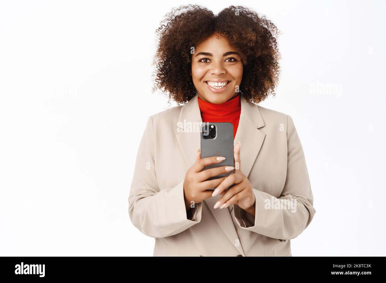 Image of smiling african american saleswoman records video on mobile phone, takes photo, shoots on smartphone, stands over white background Stock Photo