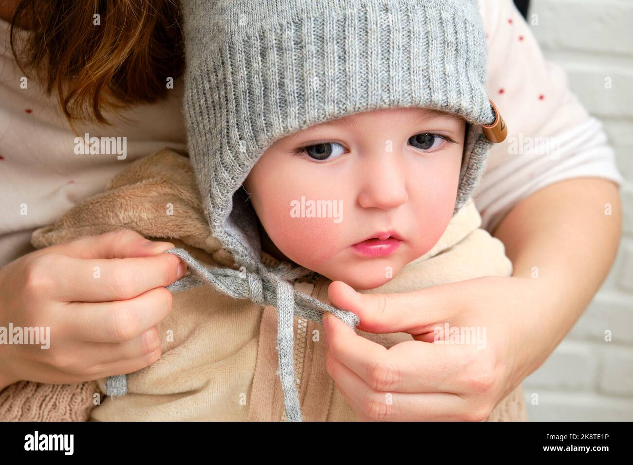 The mother puts a warm hat on the head toddler baby sitting in the home