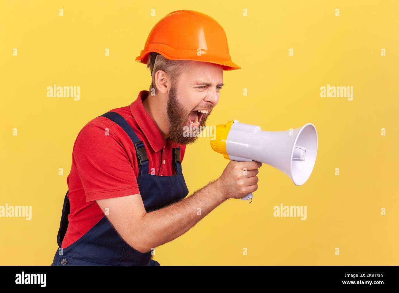 Side view of angry bearded worker wearing protective helmet and blue overalls holding megaphone and screaming with aggressive expression, protesting. Indoor studio shot isolated on yellow background. Stock Photo
