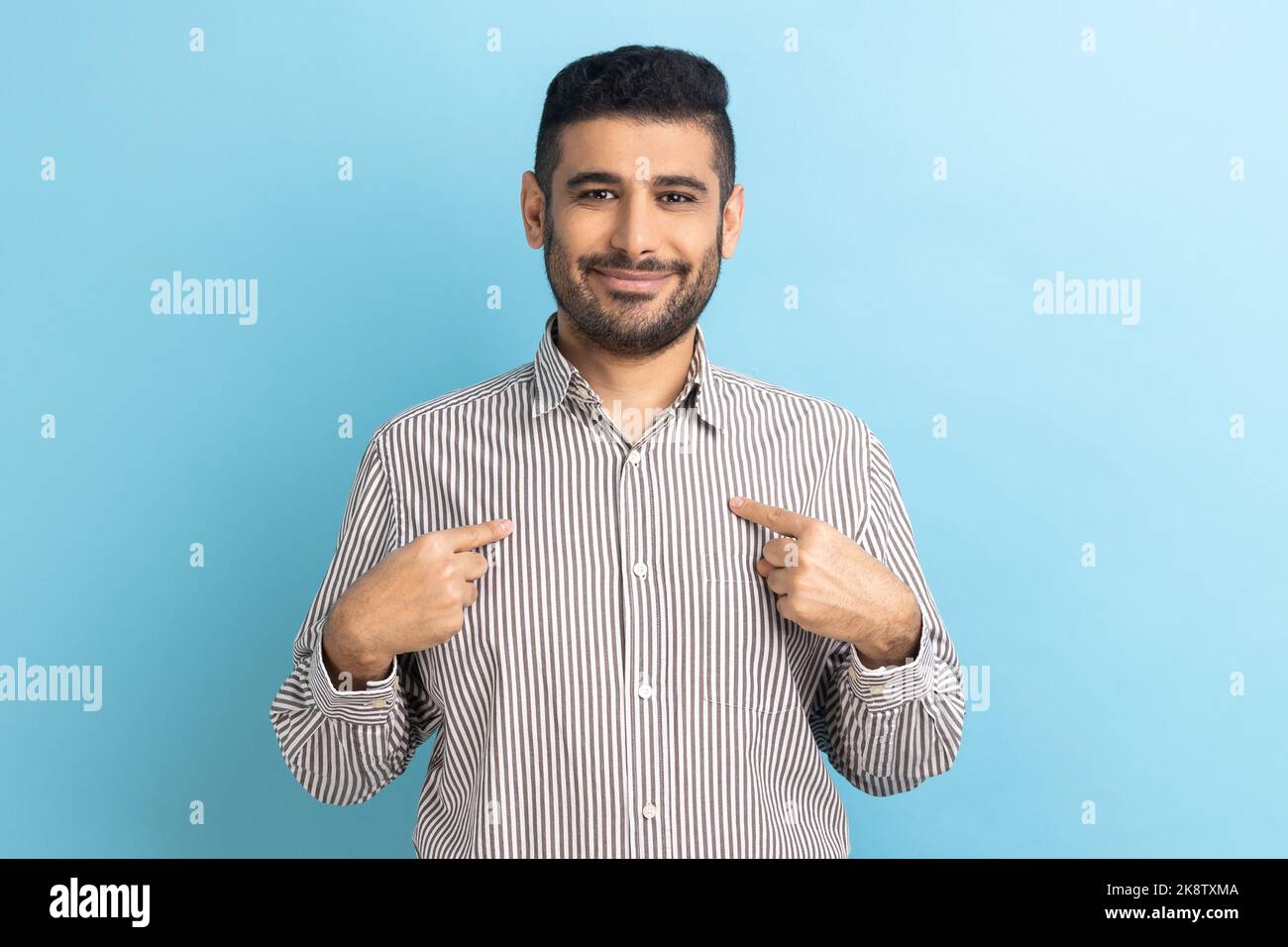 It's me. Portrait of ambitious bearded businessman with toothy smile, pointing fingers on himself, being confident, wearing striped shirt. Indoor studio shot isolated on blue background. Stock Photo