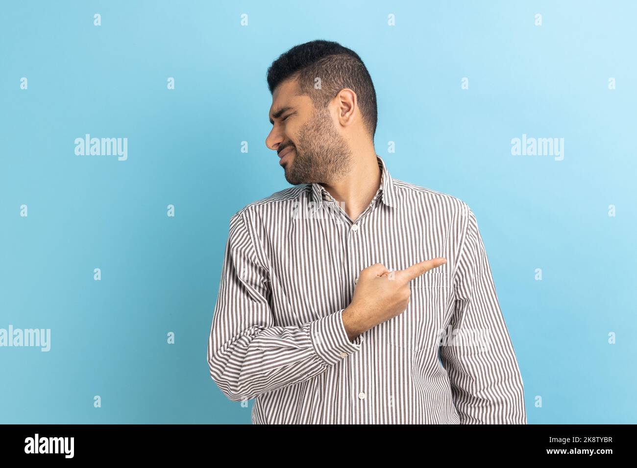 Get out. Portrait of serious angry businessman pointing finger, sneak male blaming another people, turning face, wearing striped shirt. Indoor studio shot isolated on blue background. Stock Photo