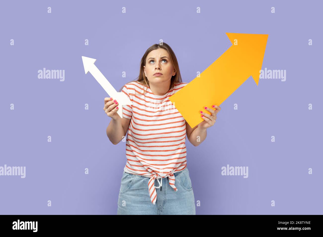 Portrait of confused puzzled blond woman wearing striped T-shirt holding two arrows indicating to different sizes, looking up and shrugging. Indoor studio shot isolated on purple background. Stock Photo