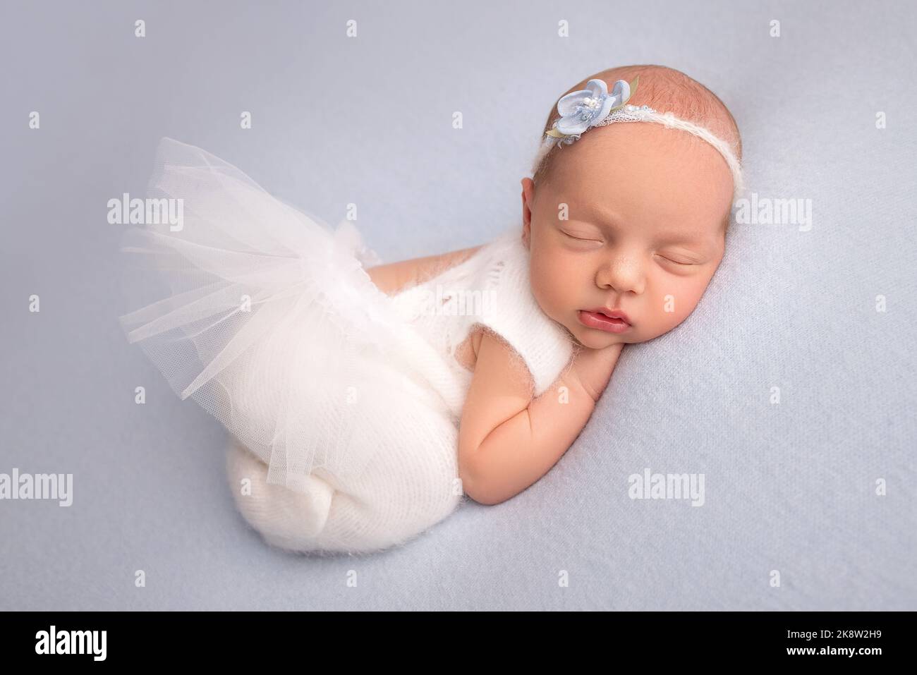 Sleeping newborn girl in the first days of life in a white ballet dress with a white bandage and a blue flower. Stock Photo