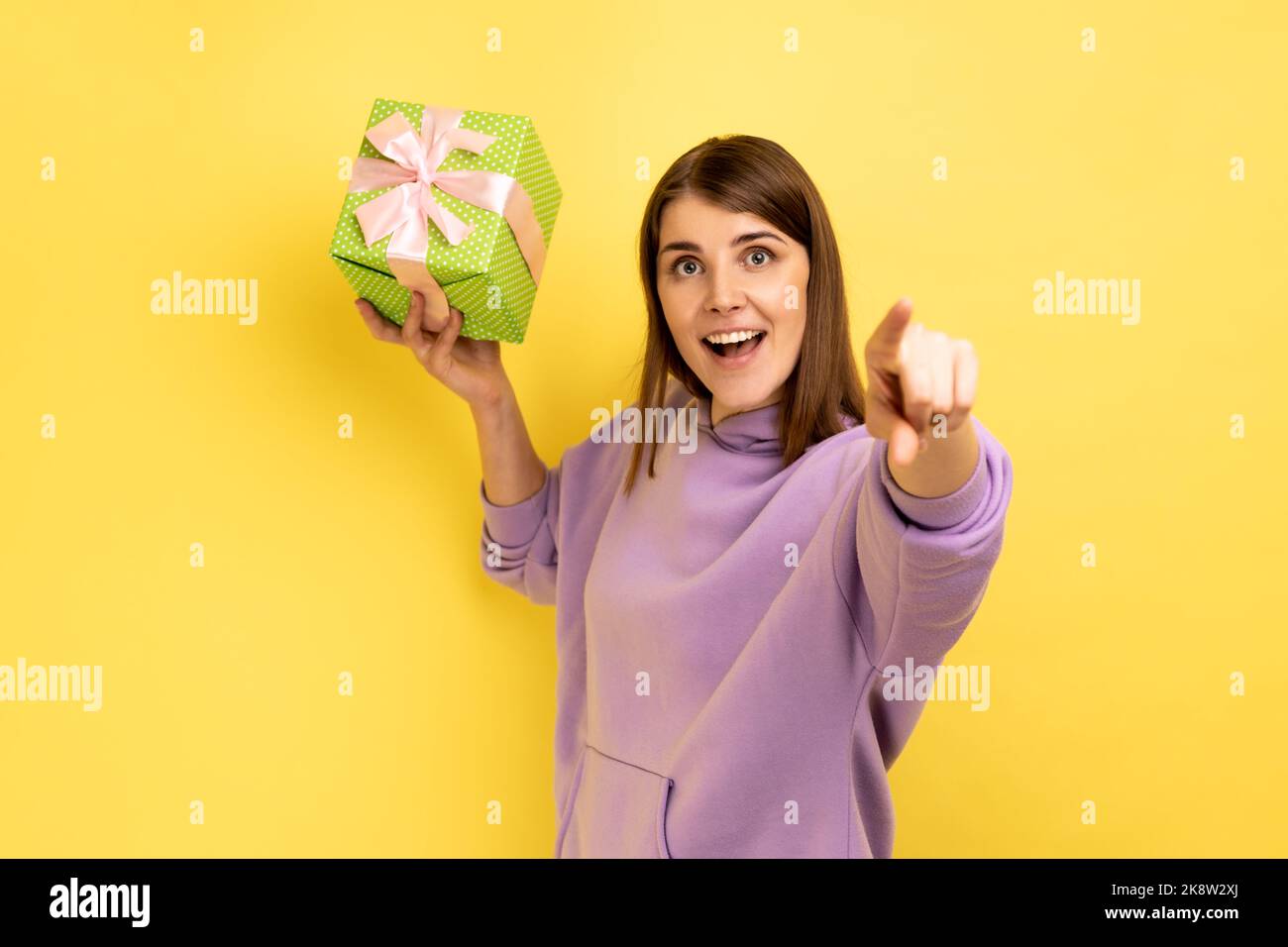 Happy excited woman throwing present box, having happy look, celebrating holiday, congratulating and pointing to camera, wearing purple hoodie. Indoor studio shot isolated on yellow background. Stock Photo