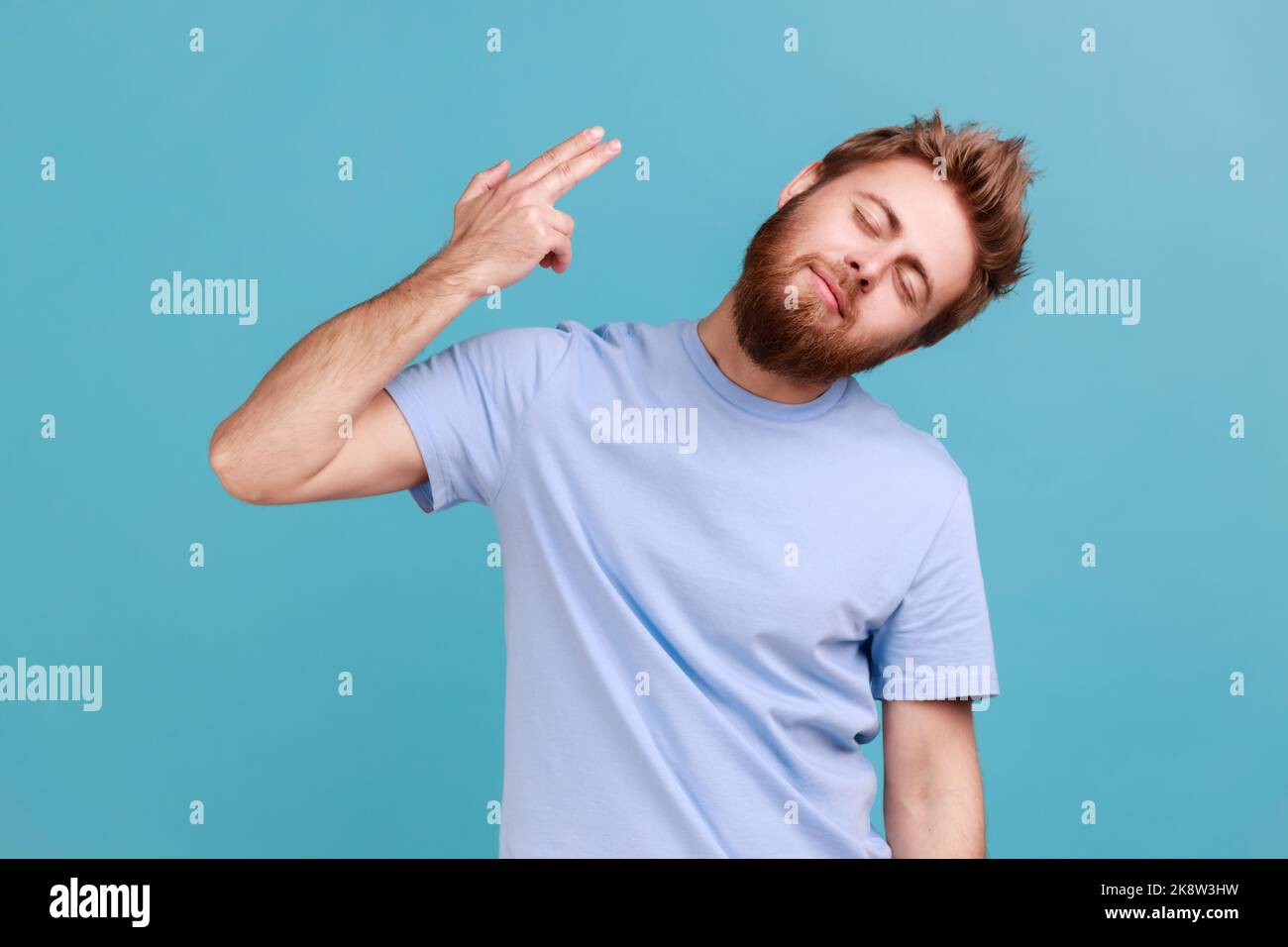 Portrait of tired depressed bearded man holding fingers near temple pretending holding gun, going to make shot, faced with problems and crisis. Indoor studio shot isolated on blue background. Stock Photo