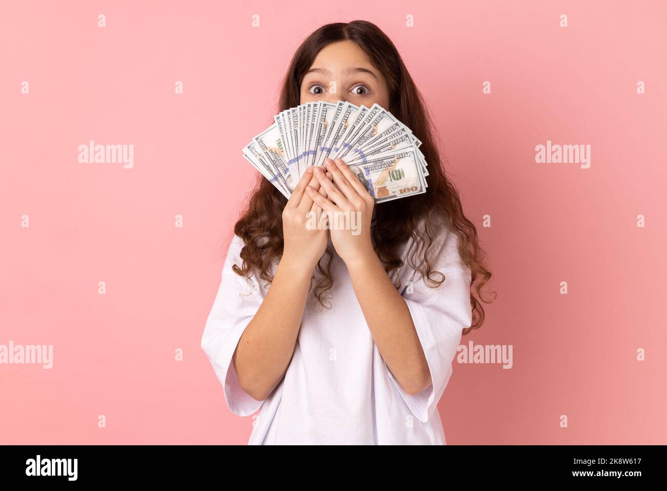 Portrait of surprised little girl wearing white T-shirt covering half of face with big fan of dollar banknotes, looking at camera with big eyes. Indoor studio shot isolated on pink background. Stock Photo