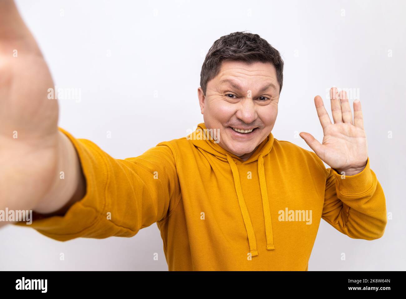 Portrait of positive handsome man making selfie or streaming, waving hand POV, point of view of photo, wearing urban style hoodie. Indoor studio shot isolated on white background. Stock Photo