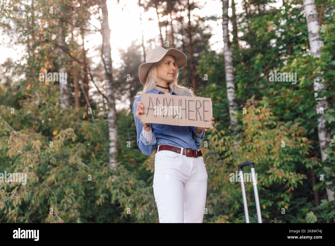 Woman wait passing car in forest standing with suitcase and cardboard