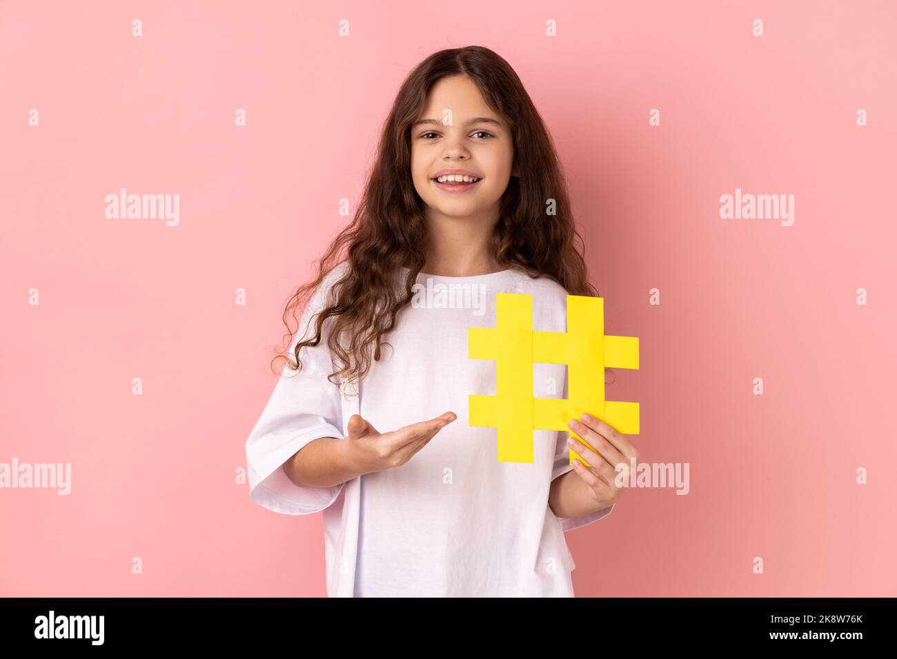 Portrait of little girl wearing white T-shirt presenting big yellow hashtag symbol and smiling at camera, blogging and child content. Indoor studio shot isolated on pink background. Stock Photo
