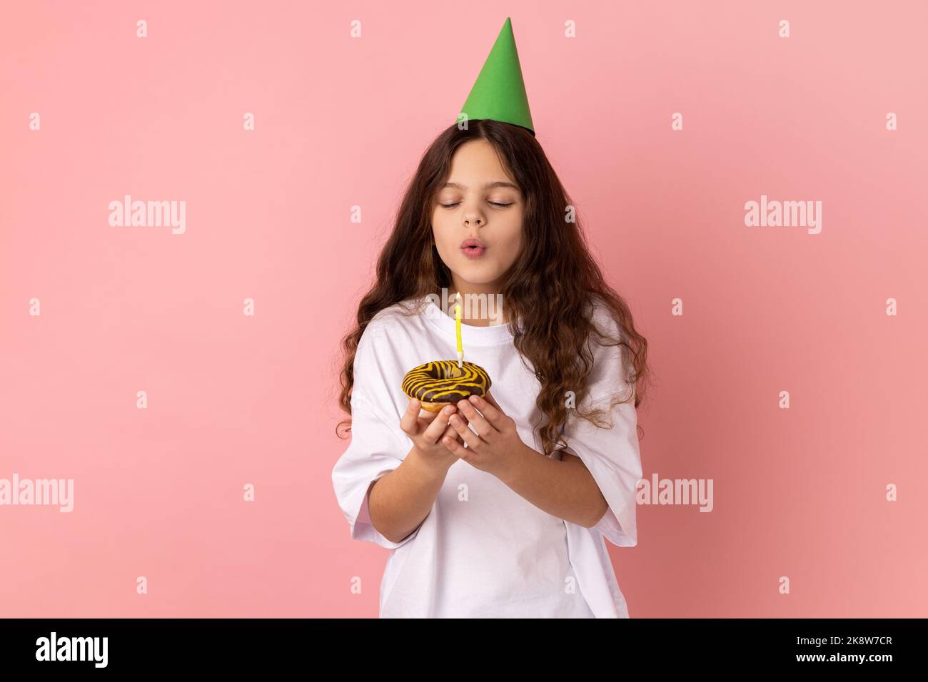 Cute charming little girl in green party cone on head holding cake and blowing candle, making wish, expressing happiness, keeps eyes closed. Indoor studio shot isolated on pink background. Stock Photo