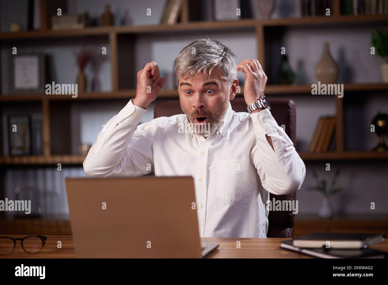 Happy businessman surprised by the good news and making yes gesture while looking at laptop sitting at office. Trader enjoying success, celebrating Stock Photo