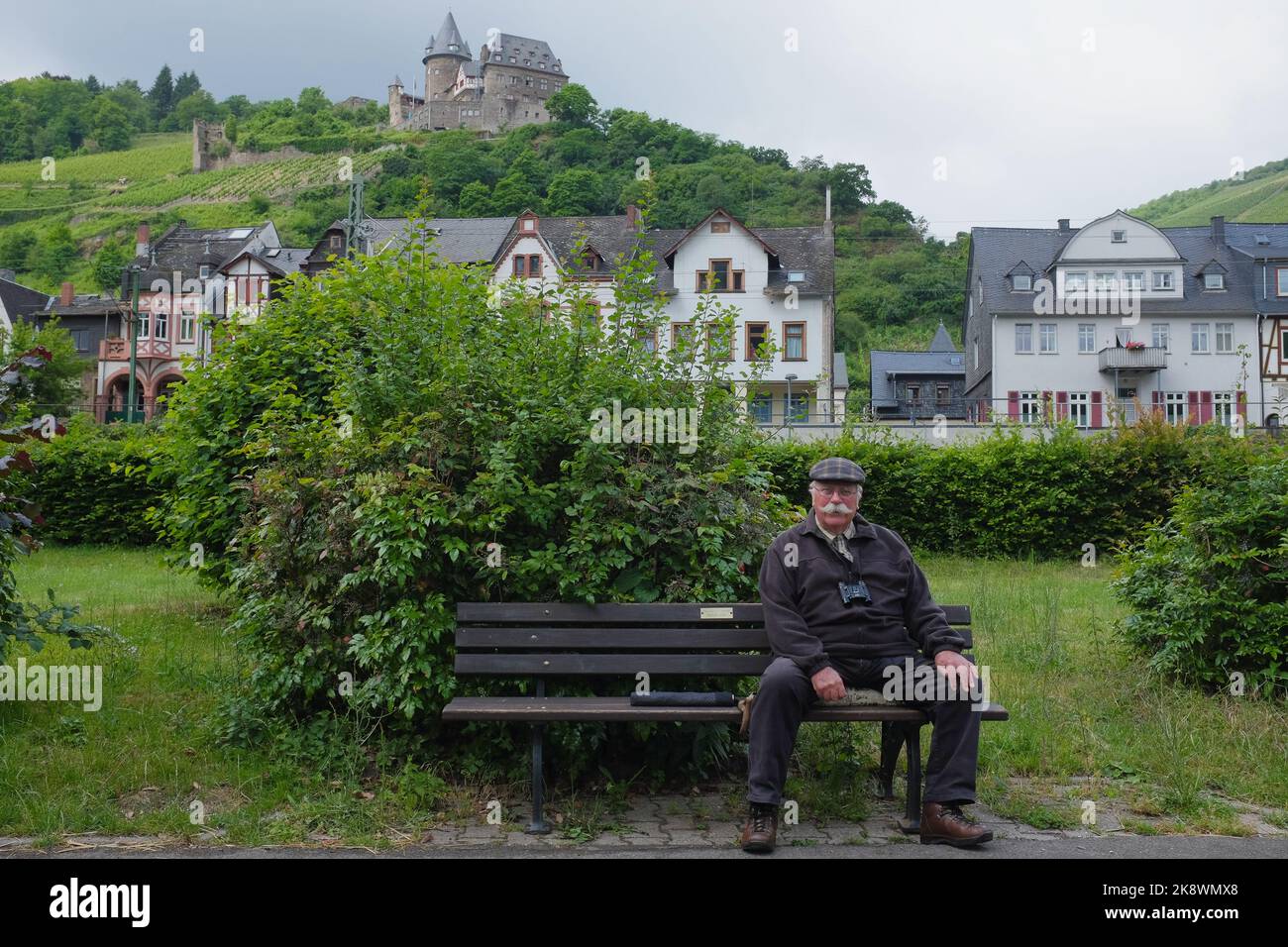 Old man with flat cap, handlebar moustache, and binoculars sits on bench in quaint Bacharach, Germany. Healthy agile senior travels on his retirement. Stock Photo