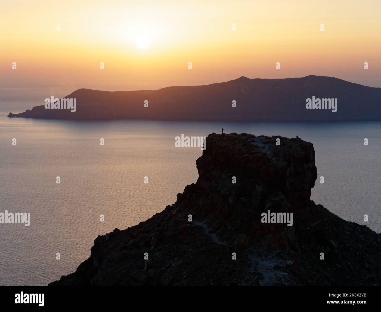 A hiker stands atop Skaros Rock foreground as the sun sets over Thirasia, on the Greek Aegean Cyclades Island of Santorini. Stock Photo