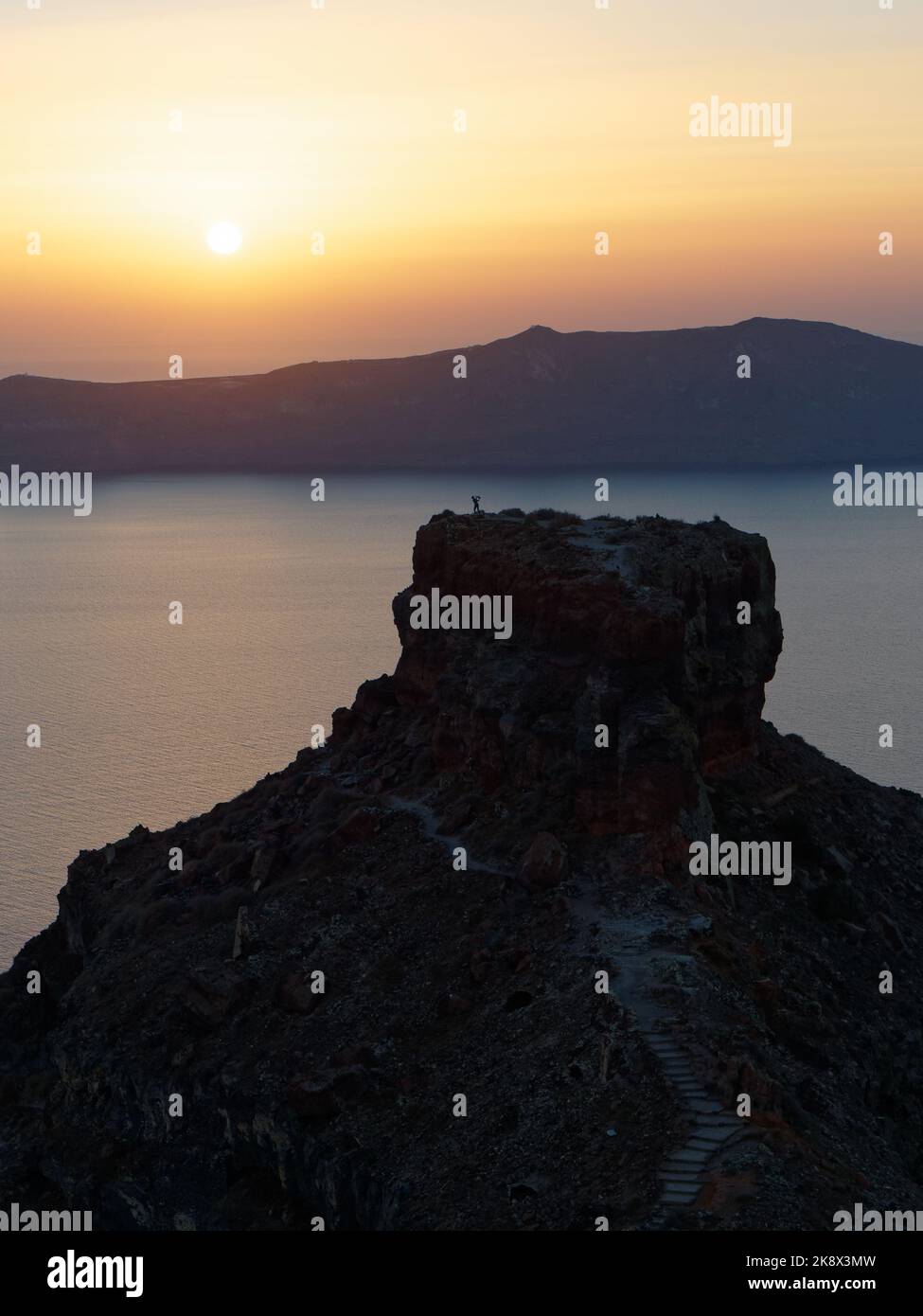 A hiker stands atop Skaros Rock foreground as the sun sets over Thirasia, on the Greek Aegean Cyclades Island of Santorini. Stock Photo