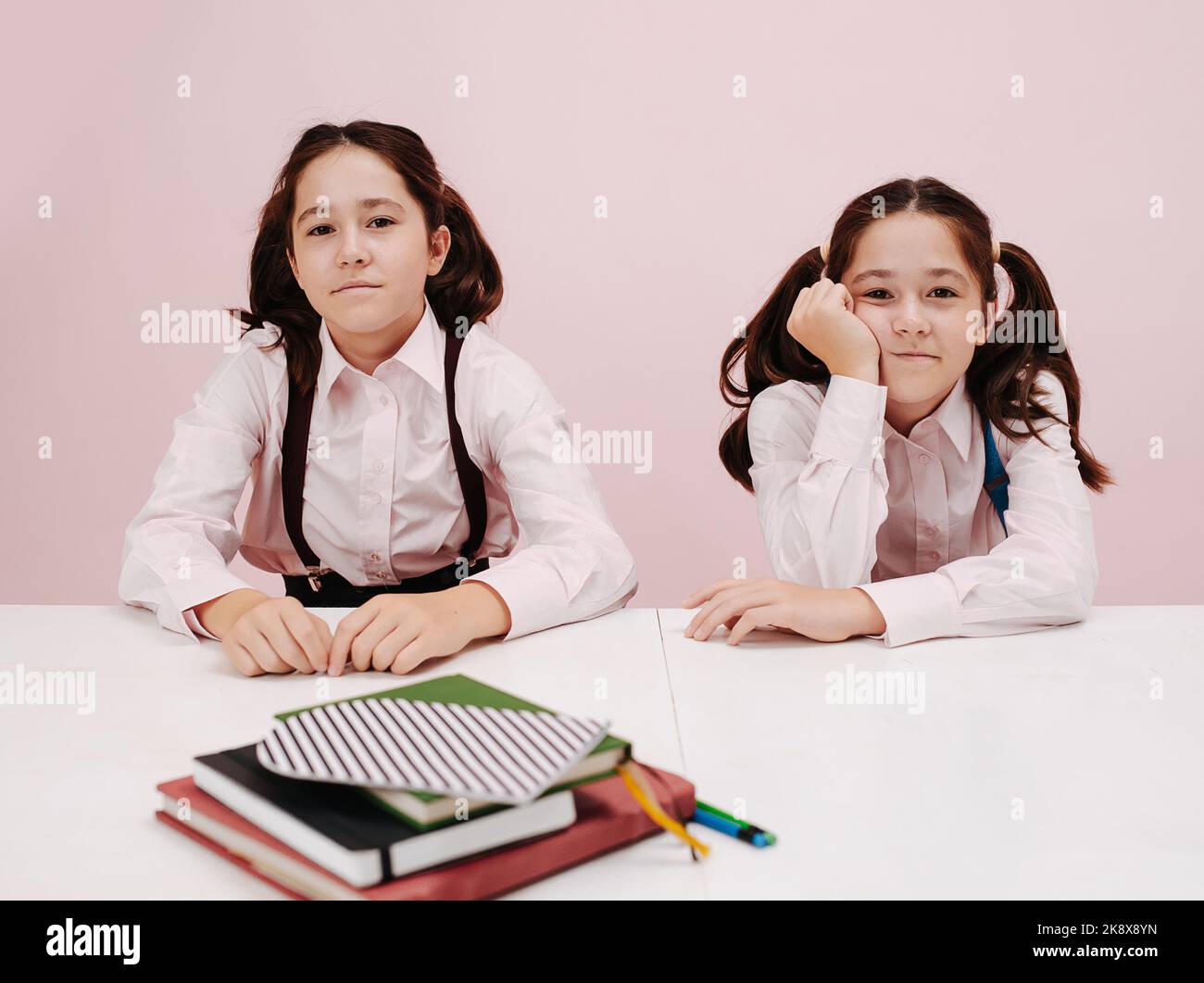 Slightly bored and tired twin schoolgirls sitting behind the desk. They are wearig twintails, shirts and suspenders. over pink background. Stock Photo