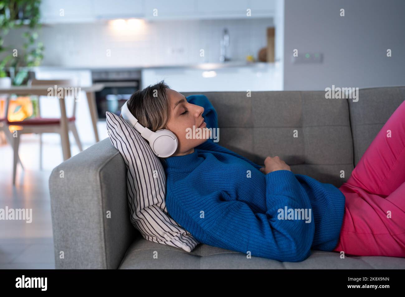 Young woman in white headphones enjoys listening to calm music and rests on comfortable sofa Stock Photo