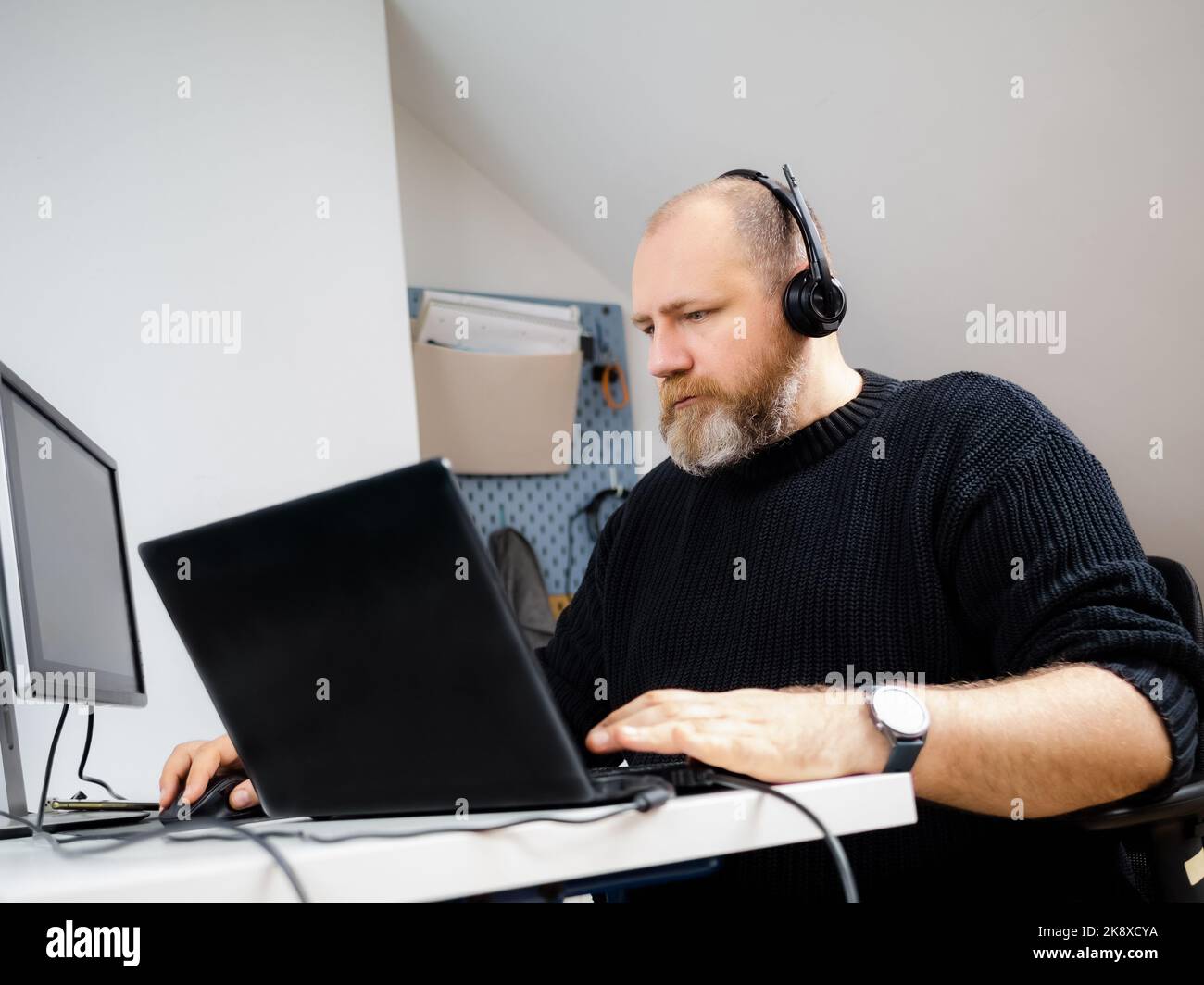 Adult bearded man remote working on laptop and using two monitors and headphones from home Stock Photo