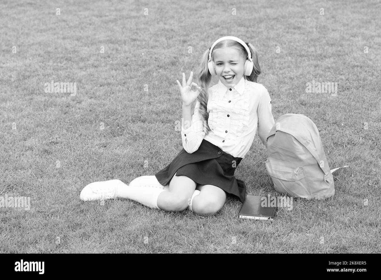 Happy girl child in school uniform show OK ring gesture sitting on green grass, satisfaction Stock Photo