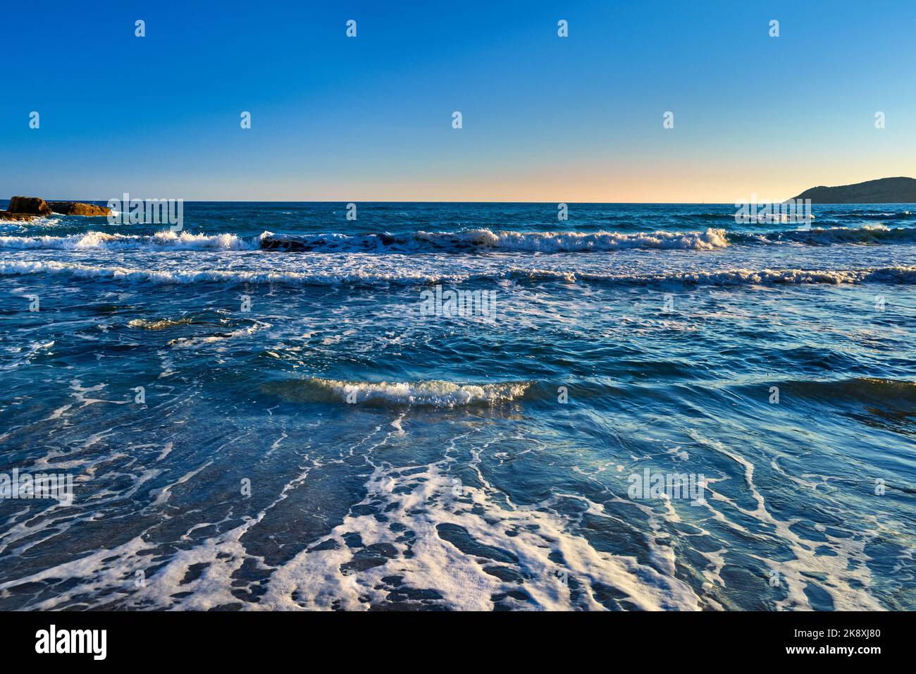 Beautiful seascape of low tidal waves, clear sunset sky, hazy hills. Clear blue and azure waters approaching with little white foam.  Stock Photo