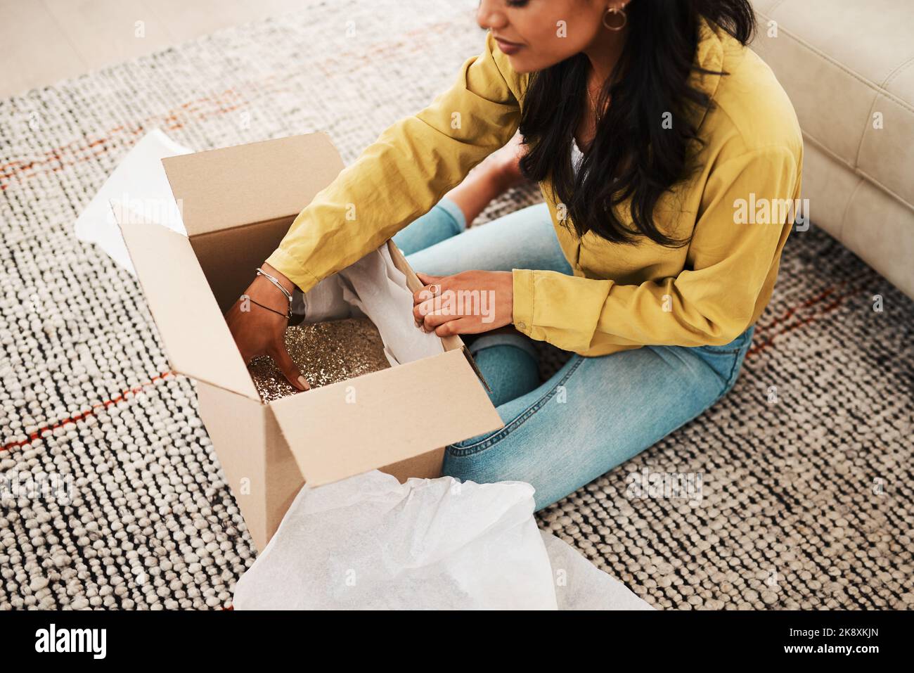 Come and take a look. an unrecognizable businesswoman sitting alone on her living room floor and opening up a mystery box. Stock Photo