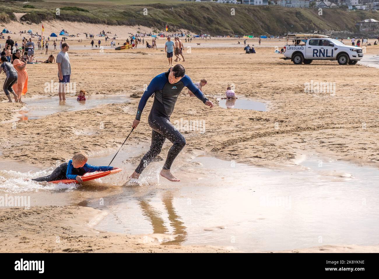 A young boy having fun lying on a body board being dragged along by his father on a busy Fistral Beach Newquay in Cornwall in the UK. Stock Photo