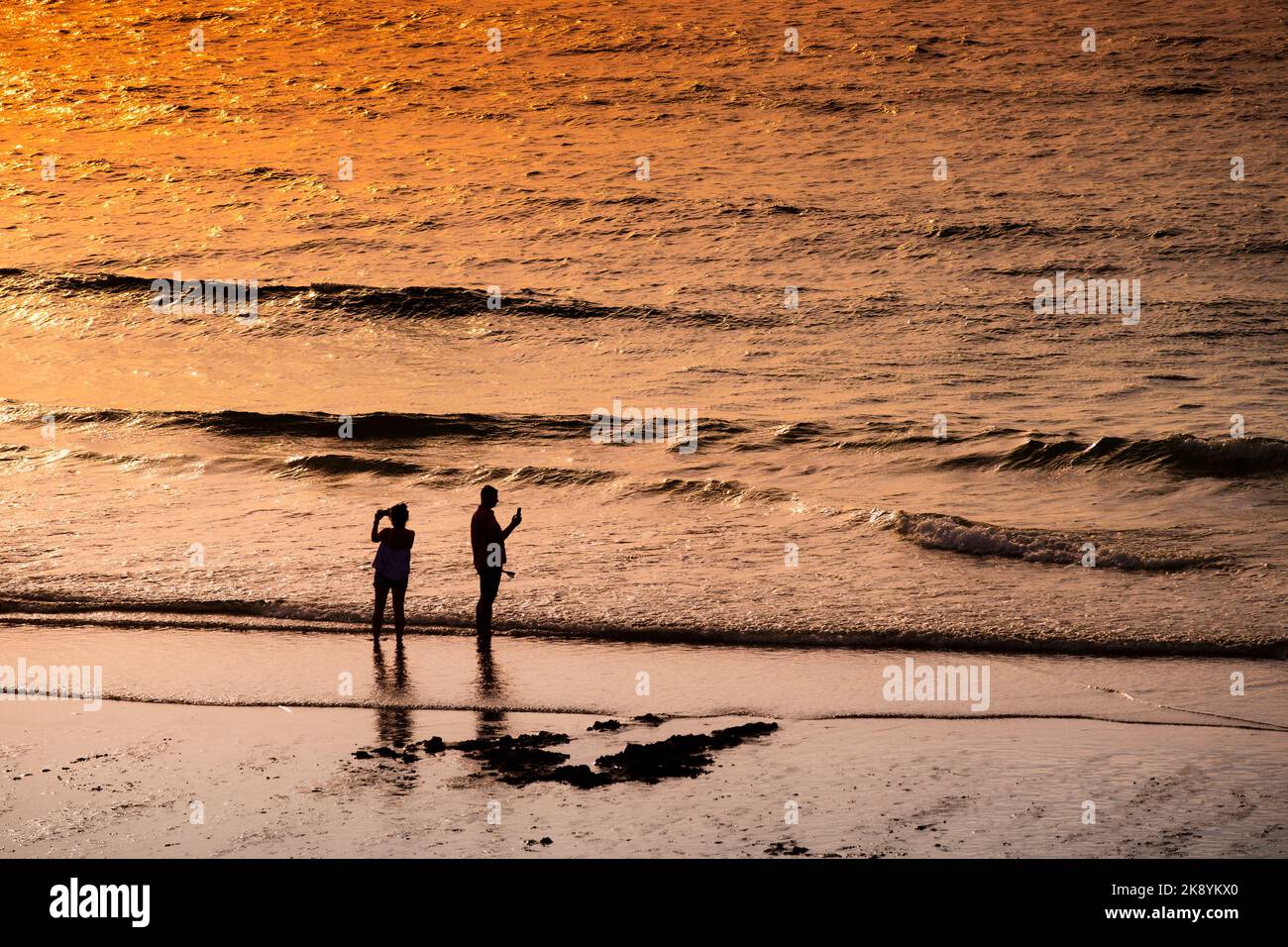 Two holidaymakers silhouetted standing on the shoreline on Fistral Beach at sunset in Newquay in Cornwall in the UK in Europe. Stock Photo