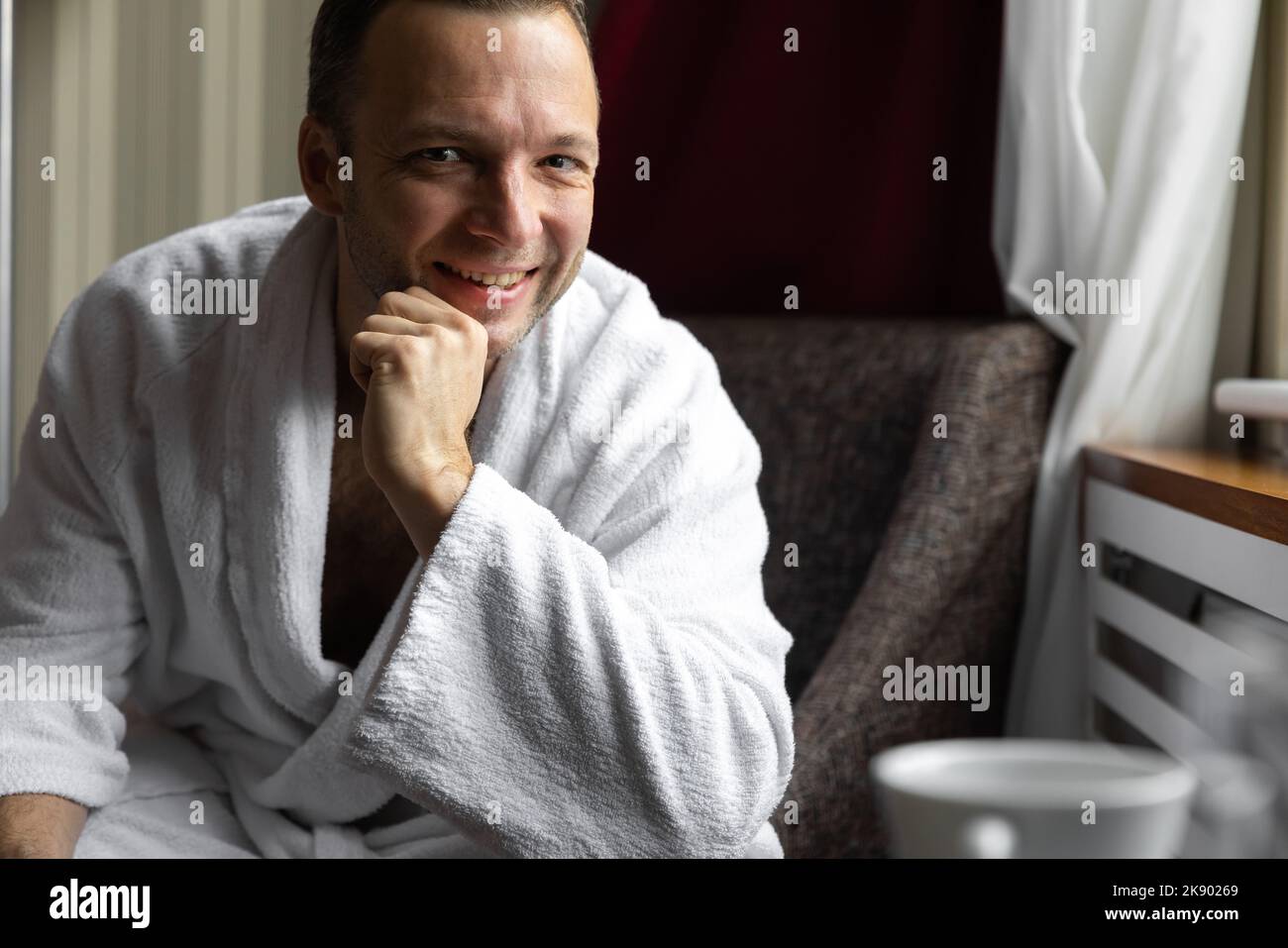 Smiling young adult Caucasian man in white bathrobe is sitting in an armchair Stock Photo