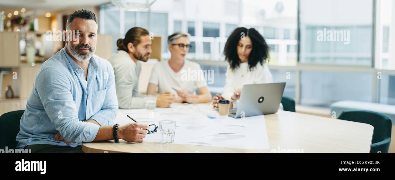 Experienced design professional looking at the camera while sitting in a meeting with his team. Group of innovative design professionals sharing creat Stock Photo