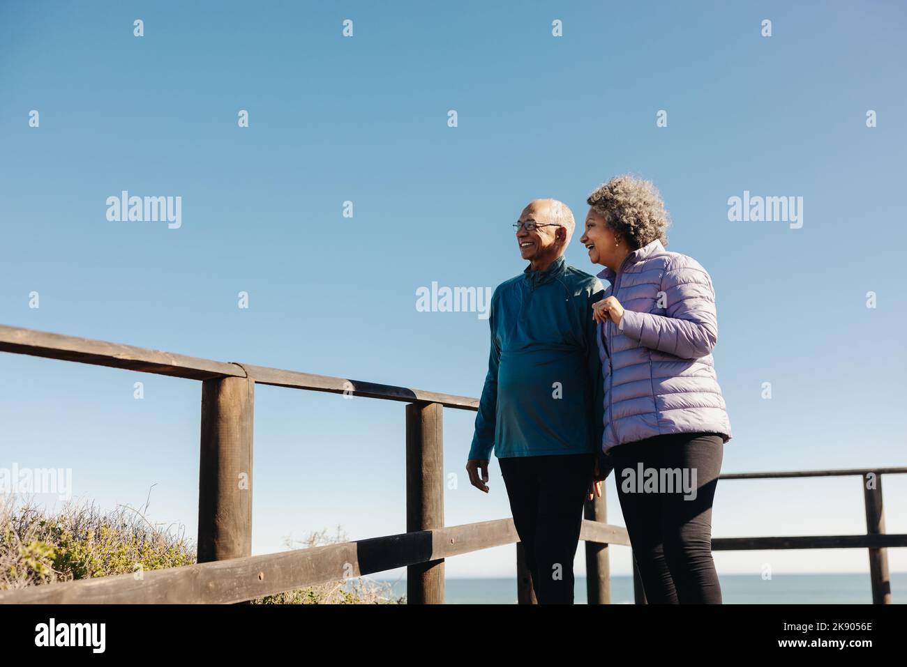 Happy senior couple looking at a refreshing view while standing on a wooden foot bridge at the ocean. Cheerful elderly couple enjoying a relaxing seas Stock Photo