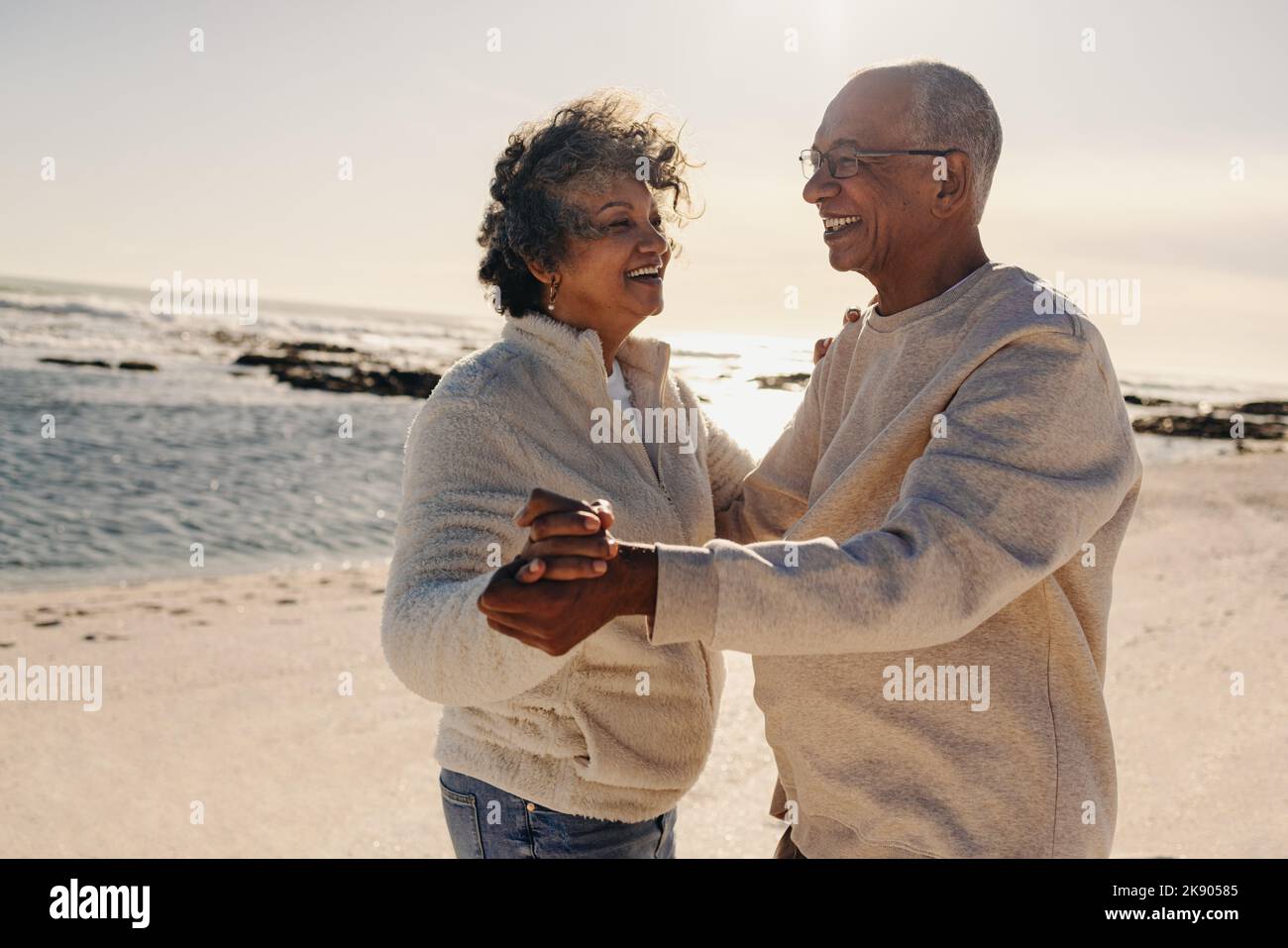 Senior couple smiling while dancing together at the beach. Happy elderly couple having a good time next to the ocean. Cheerful mature couple enjoying Stock Photo