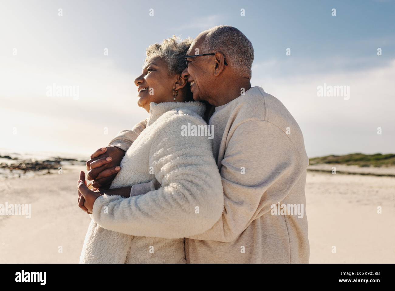 Senior couple enjoying a refreshing view of the ocean at the beach. Happy elderly couple embracing each other while standing on beach sand. Mature cou Stock Photo