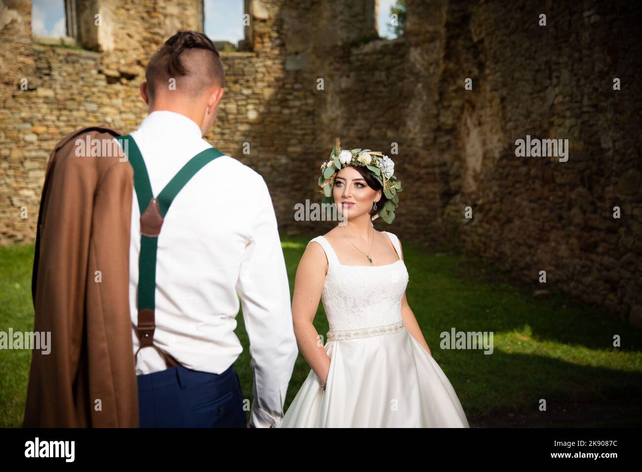 A beautiful couple in a suit and wedding dress poses for camera next to old stone walls of the castle Stock Photo