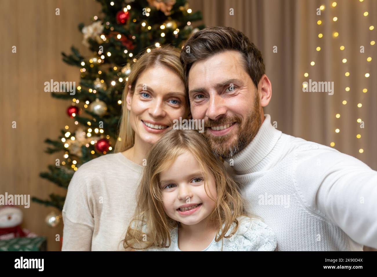 Portrait of happy family on Christmas, family man woman and daughter looking at camera and smiling, celebrating new year together at home, using phone for video call and remote communication online. Stock Photo