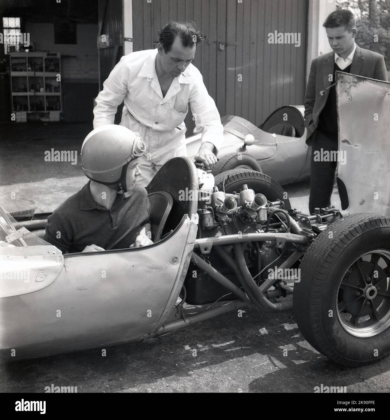 1962, historical, a man sitting in a single-seater racing car with the engine exposed with mechanic at Geoff Clarke’s Motor Racing Stables, a driving school based at the Finmere Aerodome, a disused RAF airfield near to Buckingham, England, UK. The racing school’s 1,000 cc single-seaters were rear-engined Formula Junior Coopers. Stock Photo