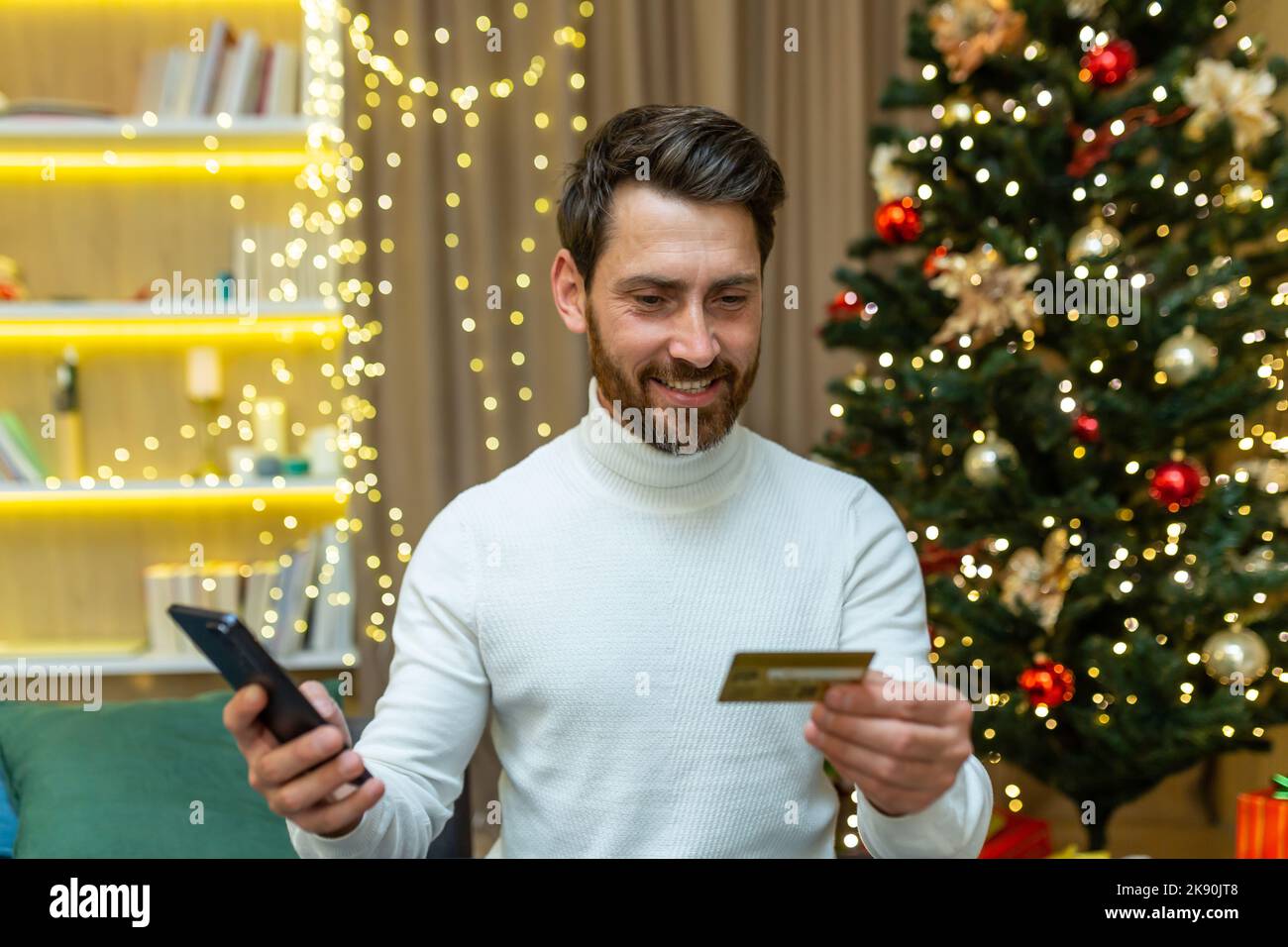 Man alone at home on Christmas celebrating new year, sitting on sofa and holding smartphone and bank credit card, happy smiling online shopping choosing gifts for friends and family remotely. Stock Photo