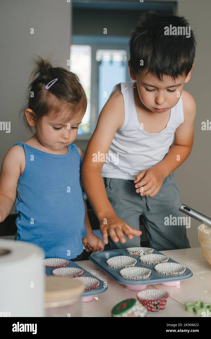 Two little caucasian kids filling the dough into cupcake silicone molds in the kitchen. Concept of confectionery cooking. Muffins making. Raw dough Stock Photo
